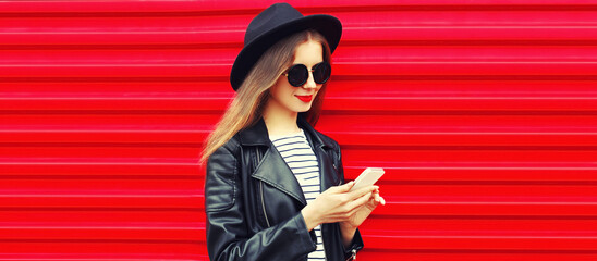 Portrait of stylish smiling woman with smartphone wearing black round hat, leather biker rock jacket on red background, blank copy space for advertising text