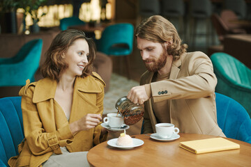 Serious handsome young man with beard sitting at table and calling colleague by phone while waiting for him in cafe