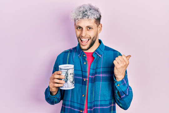 Young hispanic man with modern dyed hair holding piggy bank pointing thumb up to the side smiling happy with open mouth