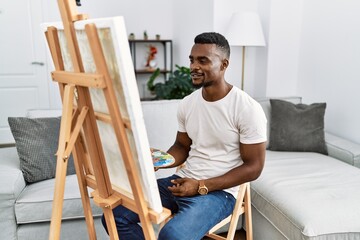 Young african man painting on canvas at home looking to side, relax profile pose with natural face and confident smile.