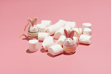 Two bright figurines of easter bunnies surrounded by white fluffy marshmallows on a pink surface, a close up