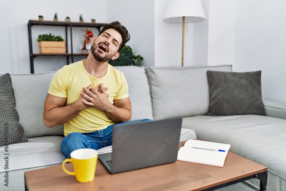 Poster Young man with beard using laptop at home smiling with hands on chest with closed eyes and grateful gesture on face. health concept.