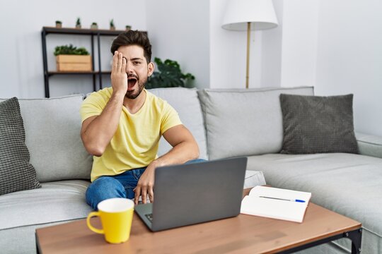 Young man with beard using laptop at home covering one eye with hand, confident smile on face and surprise emotion.