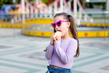 portrait of a child girl in an amusement park in the summer eating ice cream near the carousels and smiling with happiness in sunglasses, the concept of weekends and school holidays