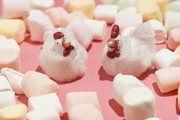 Two white chicken figurines surrounded by colorful fluffy marshmallows on a pink surface, a close up
