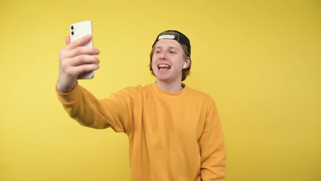 Joyful guy in a cap and casual clothes takes a selfie on the smartphone camera on a yellow background and laughs and makes a video call