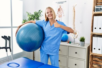 Young caucasian physiotherapist woman holding pilates ball smiling happy and positive, thumb up...
