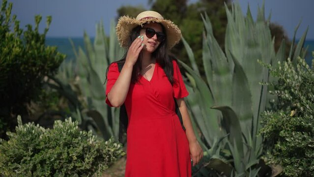 Beautiful young Caucasian woman in red dress and sunglasses posing at background of beautiful Cyprus nature. Portrait of confident female tourist looking at camera smiling standing in sunshine