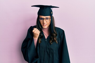 Young hispanic woman wearing graduation cap and ceremony robe angry and mad raising fist frustrated and furious while shouting with anger. rage and aggressive concept.