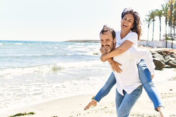 Middle age hispanic man smiling happy holding woman on his back at the beach.
