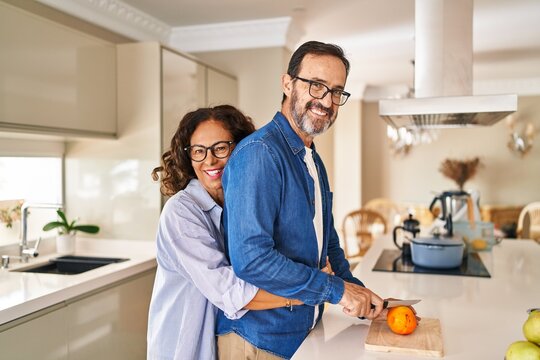Middle age hispanic couple hugging each other cutting orange at kitchen