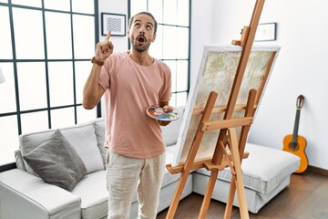 Young hispanic man with beard painting on canvas at home pointing finger up with successful idea. exited and happy. number one.