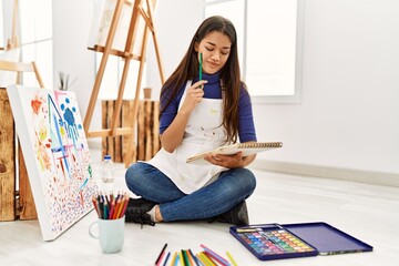 Young latin woman with doubt expression drawing on notebook at art studio