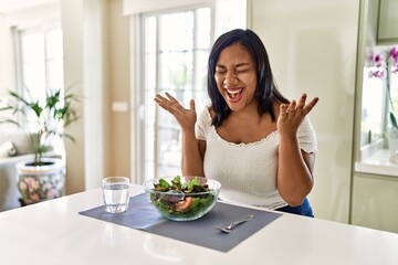Obraz na płótnie Canvas Young hispanic woman eating healthy salad at home celebrating mad and crazy for success with arms raised and closed eyes screaming excited. winner concept