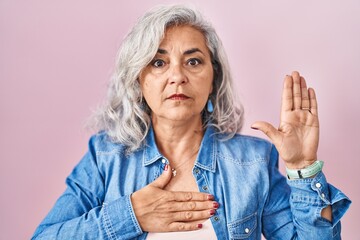 Middle age woman with grey hair standing over pink background swearing with hand on chest and open...