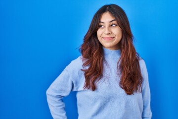 Hispanic young woman standing over blue background smiling looking to the side and staring away thinking.