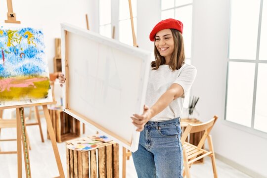 Young hispanic artist woman wearing french beret holding canvas at art studio.
