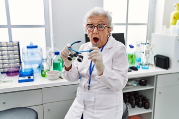 Senior woman with grey hair working at scientist laboratory using magnifying glasses afraid and...