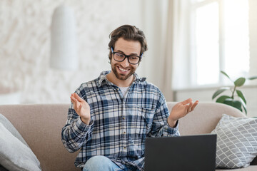 Cheerful young caucasian man teacher with stubble in glasses sits on sofa with laptop and...