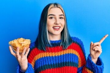 Young modern girl holding potato chips smiling happy pointing with hand and finger to the side