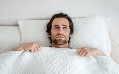 Unhappy millennial caucasian man with stubble wake up, feeling bad, lying on white bed in bedroom...