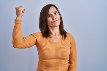 Middle age brunette woman standing wearing orange sweater angry and mad raising fist frustrated and...