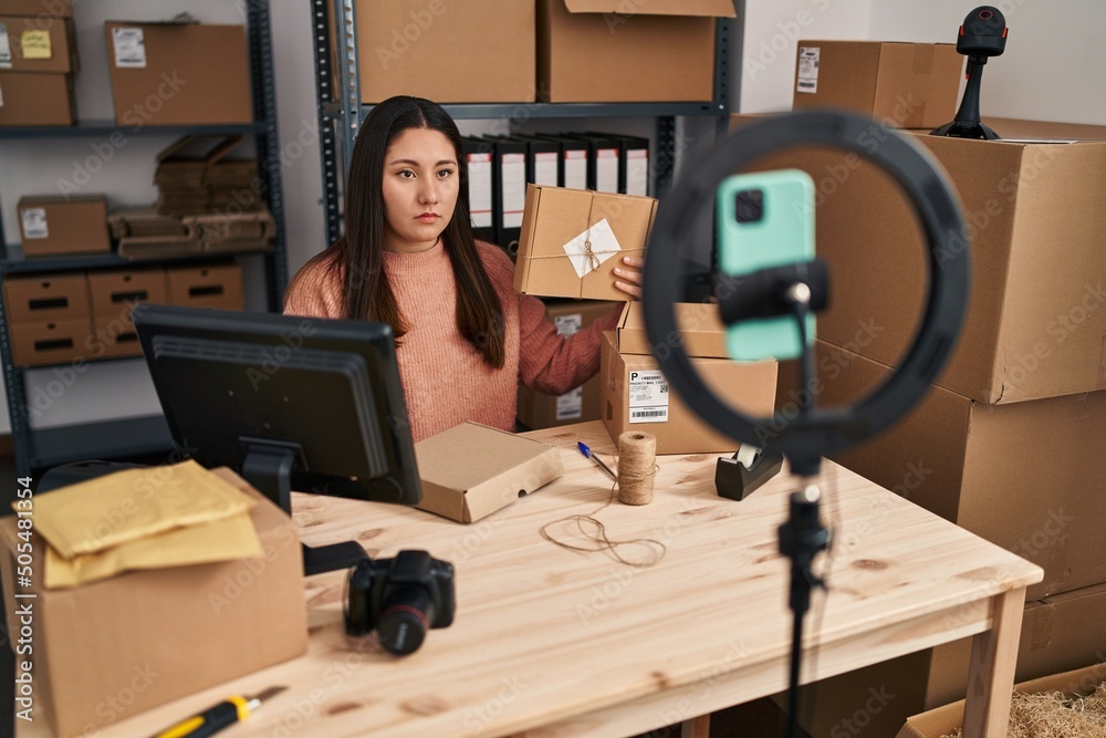 Sticker Young latin woman working at small business ecommerce doing video thinking attitude and sober expression looking self confident