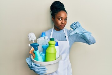 African american woman with braided hair wearing apron holding cleaning products with angry face, negative sign showing dislike with thumbs down, rejection concept