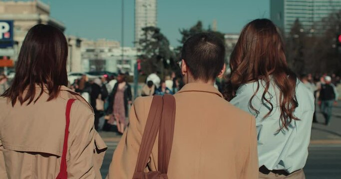 Three young women in autumn clothes stand together at street light waiting to cross on sunny windy day slow motion.