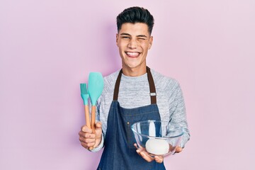 Young hispanic man holding bread dough and cooking tools winking looking at the camera with sexy...