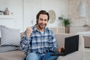 Glad young caucasian man with stubble sits on sofa make victory gesture, sits on sofa with laptop...