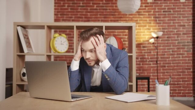 A young man at his workplace is typing on a laptop, then thoughtfully clutches his head, rubs his temples and continues to type. He looks very tense and puzzled. Slow motion 4k footage