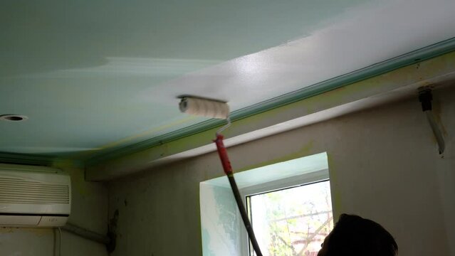 
A man makes repairs inside the house, painting the ceiling with a long stick with a roller. Ceiling covered with white paint. Water emulsion coating, renovation of an old renovation with acrylic