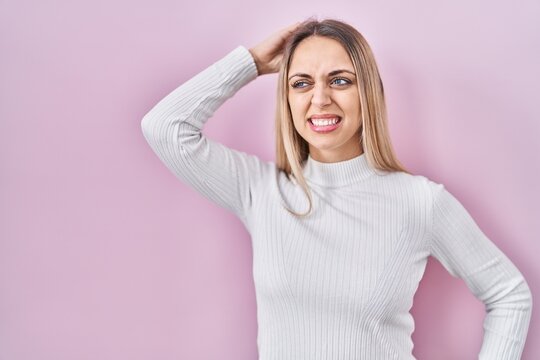 Young blonde woman wearing white sweater over pink background confuse and wonder about question. uncertain with doubt, thinking with hand on head. pensive concept.