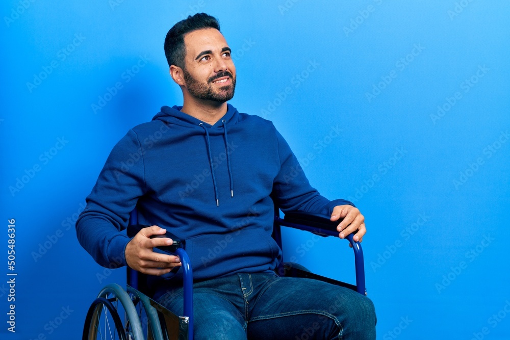 Wall mural Handsome hispanic man with beard sitting on wheelchair looking away to side with smile on face, natural expression. laughing confident.