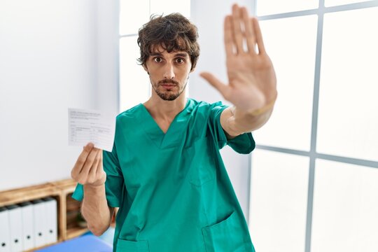 Young hispanic doctor man holding covid record card doing stop sing with palm of the hand. warning expression with negative and serious gesture on the face.