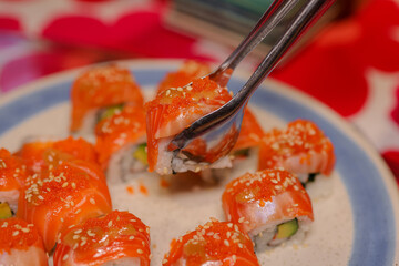 Sushi Delight: A close-up shot captures a hand using metal tongs to delicately pick up a vibrant,...