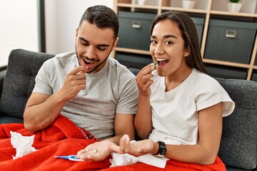Young latin ill couple eating pills sitting on the sofa at home.