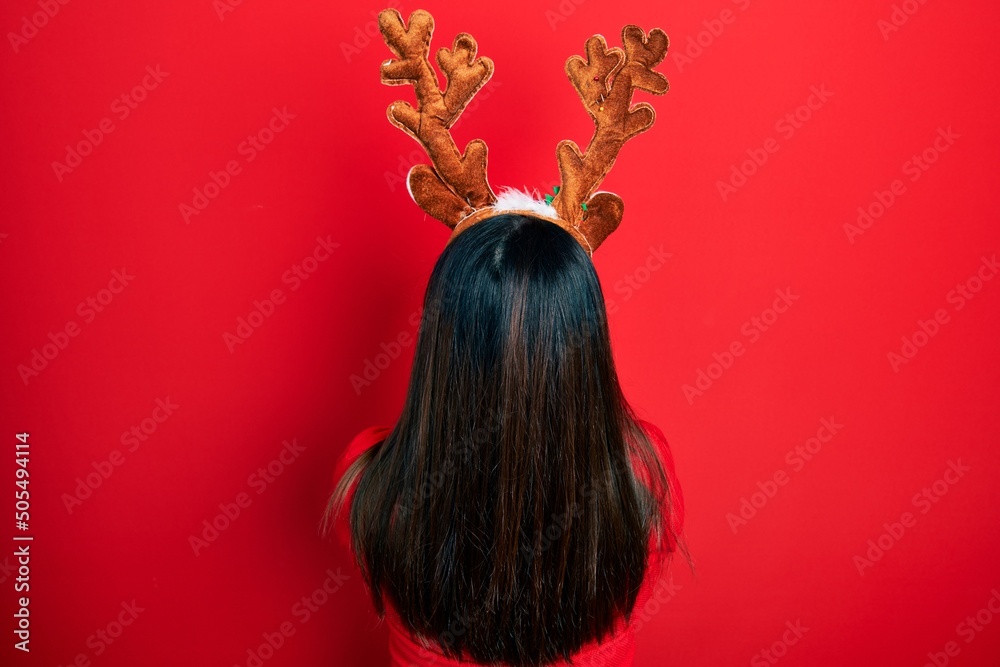 Poster young hispanic woman wearing deer christmas hat and red nose standing backwards looking away with cr