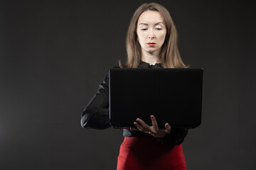 Serious woman using laptop. The businesswoman stands and looks at the laptop screen. A girl with a laptop on a dark background. Place for text. The use of electronic gadgets.