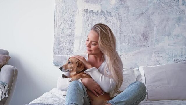 Elegant beautiful blonde woman simple clothes plays with cute adorable brown dog dachshund in the bedroom while sitting on a cozy bed in the bedroom. Time to have fun. concept of a pet. friend dog