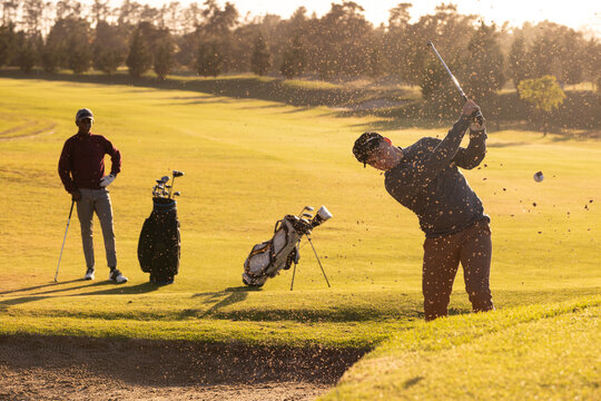 Caucasian young man hitting golf shot while playing with african american friend at course at sunset