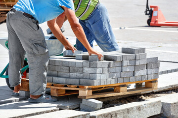 Workers unload paving slabs from a wooden pallet on a clear sunny day.
