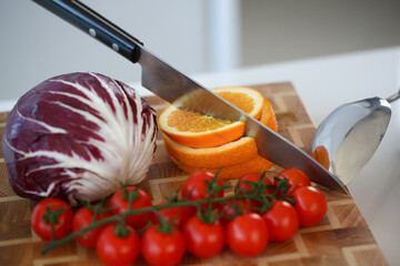 Knife in orange slices, tomatoes and kale on wooden cutting board, add ripe vegetable to salad