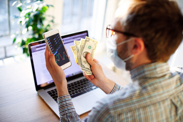 Man in mask working remotely at home with phone and laptop, counting money savings due to economic financial crisis and unemployment looking at phone with currency rates, income decrease, trading