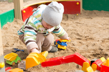 Funny little blond boy in a jacket and hat playing with plastic toy cars in the sandbox on the playground in the park. Happy childhood. Game