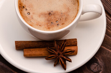 On a wooden table is a white porcelain cup with coffee, next to it on a saucer are spices.