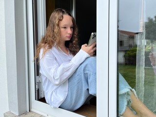 teenager sits she can be seen in the window in her hands she holds a white iPhone 13 pro max she looks 