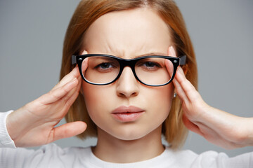 Close up of upset tired woman in eyeglasses suffering from headache and migraine, plain grey background. Touching temples, feeling stress and pain, annoyed, exhausted, people concept, horizontal