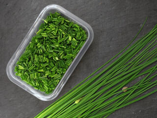 freshly cut chives in storage box next to whole bunch chives on black slate board, top view of...